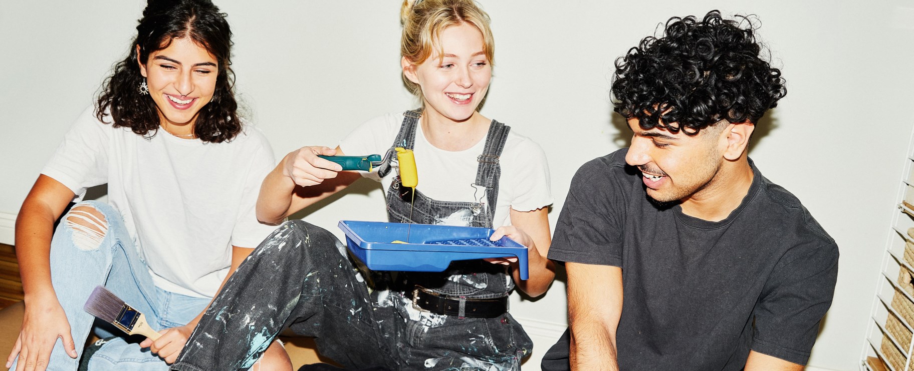 Tre glada unga personer sitter på golvet i ett rum och håller penslar och målarredskap i handen