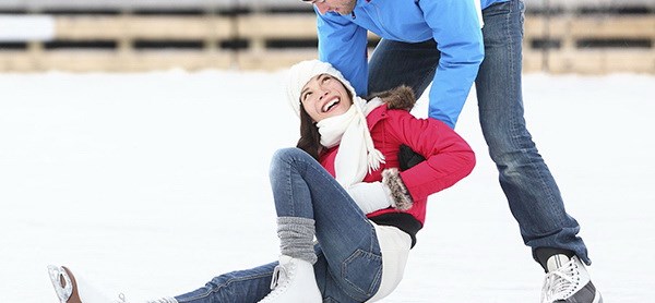 Kvinna i röd jacka har trillat på sina skridskor men får hjälp upp av man i blå jacka