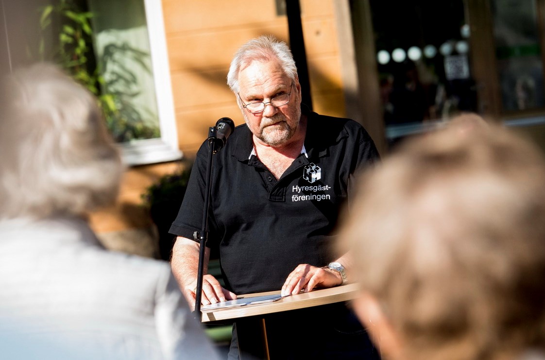 Man står vid talarstol utomhus, suddig publik i förgrunden.
