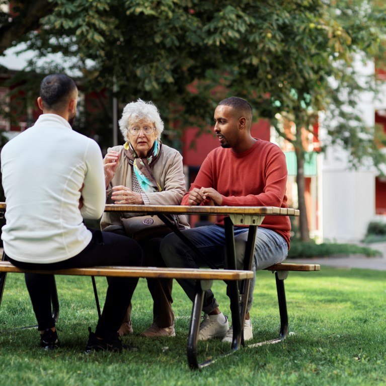 En äldre kvinna i vitt hår sitter vid ett bord på en gård tillsammans med två unga män med utländsk bakgrund.