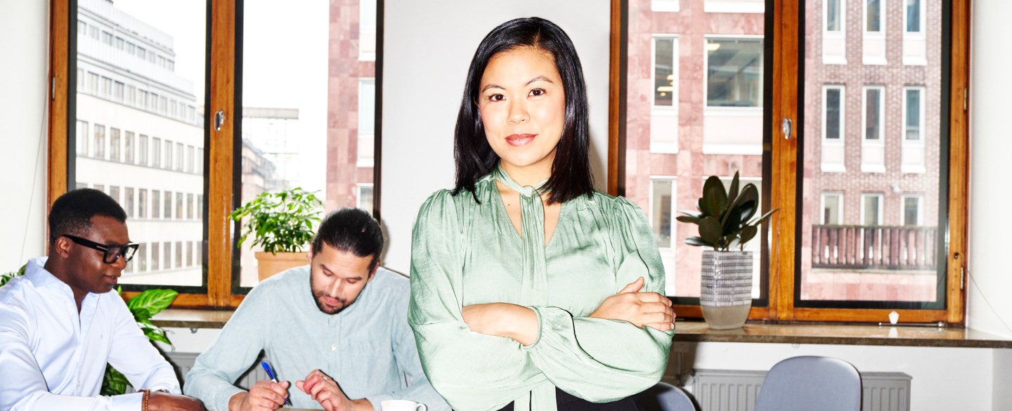 Bestämd kvinna står med händerna i kors. I bakgrunden sitter två män och skriver