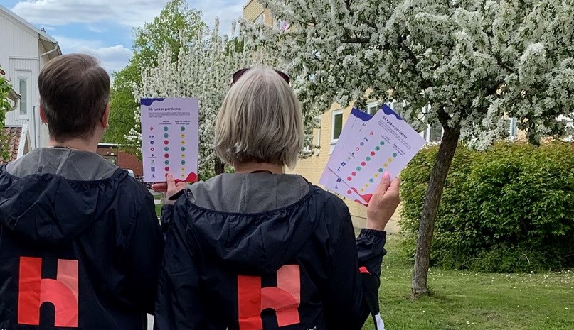 Två ryggar med logotyp, de håller kampanjblad mot blommande träd