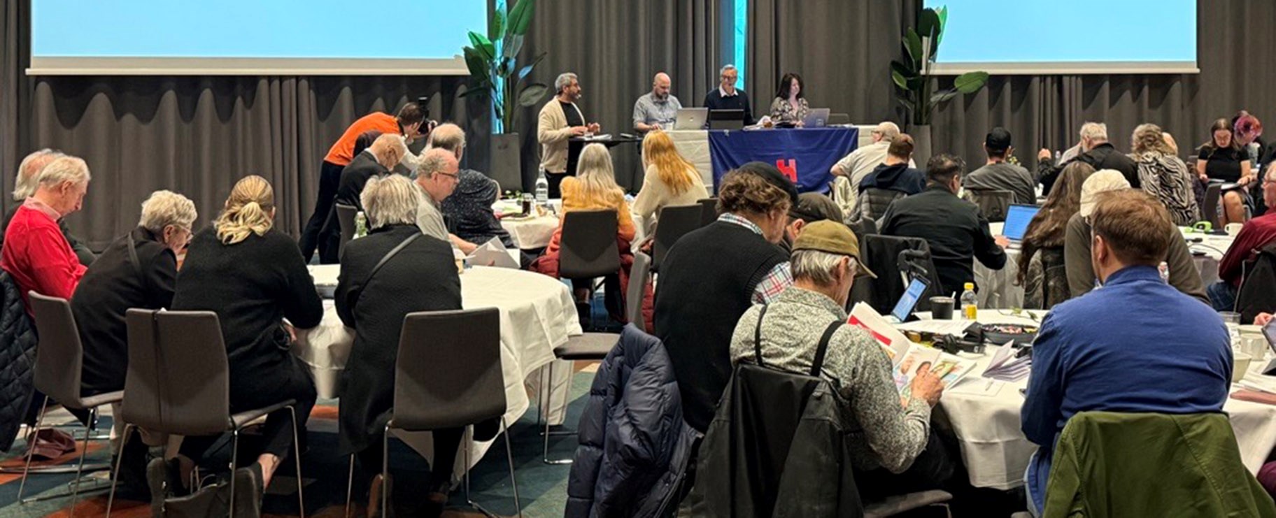 Foto från en konferenslokal med runda bord som ledamöter sitt runt. Längst fram är ett podium där presidiet sitter.