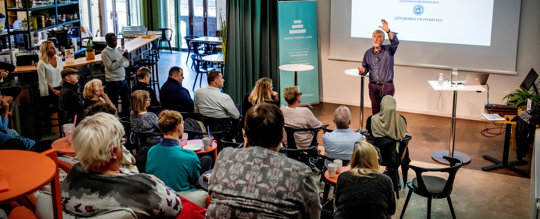 Foto från ett evenemang i vår Arena: Första Lång. Bilden är tagen från en läktare, i förgrunden ser man publik och som sitter vänd mot en föreläsare som står på scenen och pratar.