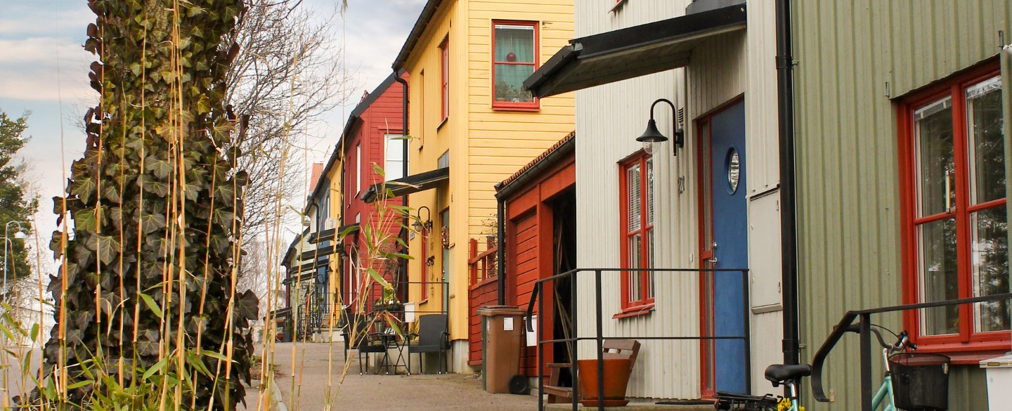 Färgglada radhus står i rad längs en trottoar, omgivna av höstväxter och träd. Husen är målade i nyanser av gult, rött, grönt och beige, med färgglada dörrar och fönsterkarmar. Framför det gröna huset står en blå cykel och en blomkruka.