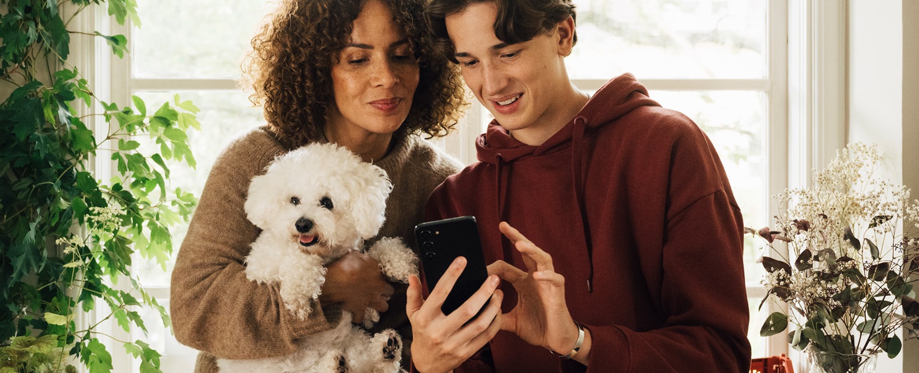 En tjej och en kille står bredvid varandra. Tjejen håller i en vit hund och killen håller fram en svart smartphone, som båda kollar på.