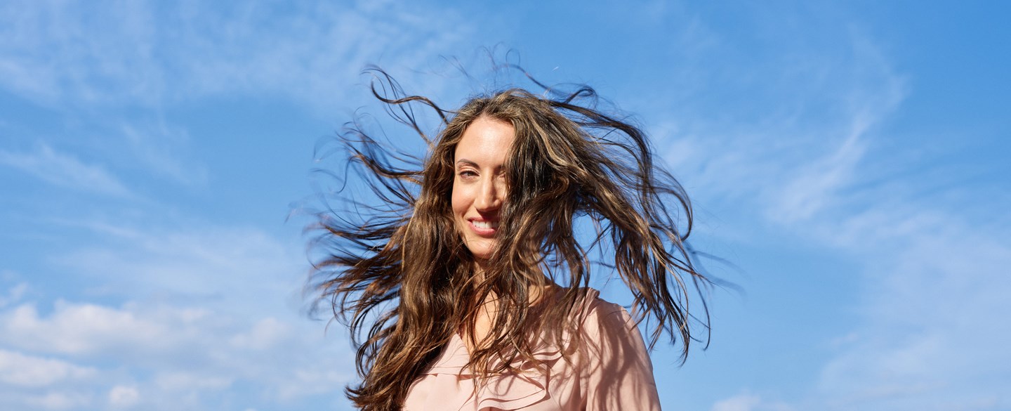 Kvinna med långt brunt hår står utomhus på en båt med en klarblå himmel i bakgrunden. Hennes hår blåser i vinden och hon bär en ljusrosa blus.