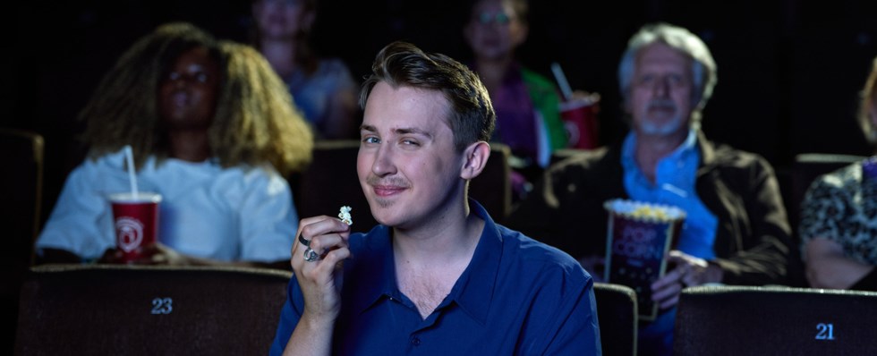 Man med kort, blont hår sitter i en biosalong och håller en skål med popcorn. Han ler och tittar in i kameran. Flera personer sitter i bakgrunden och kollar på bio.