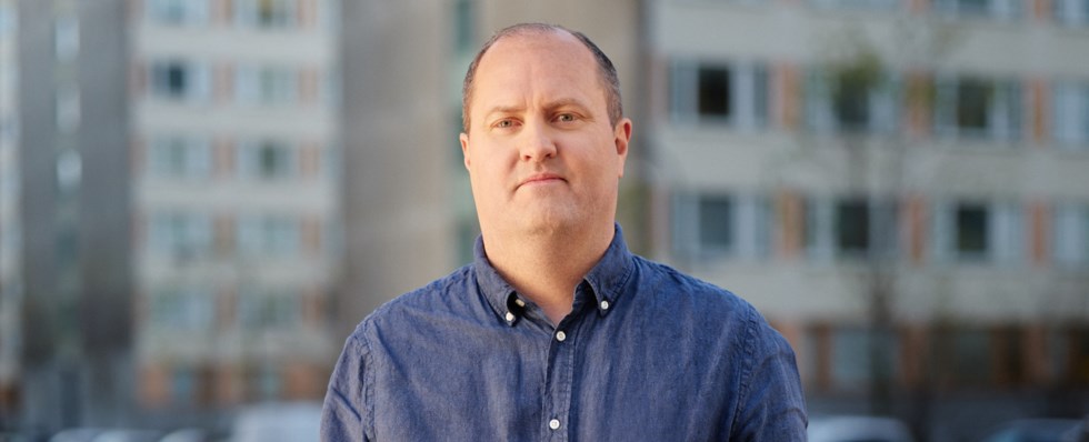 Erik Elmgren, förbundschef på Hyresgästföreningen, står utomhus framför en bakgrund av flerbostadshus. Han har på sig en blå skjorta och ser in i kameran.