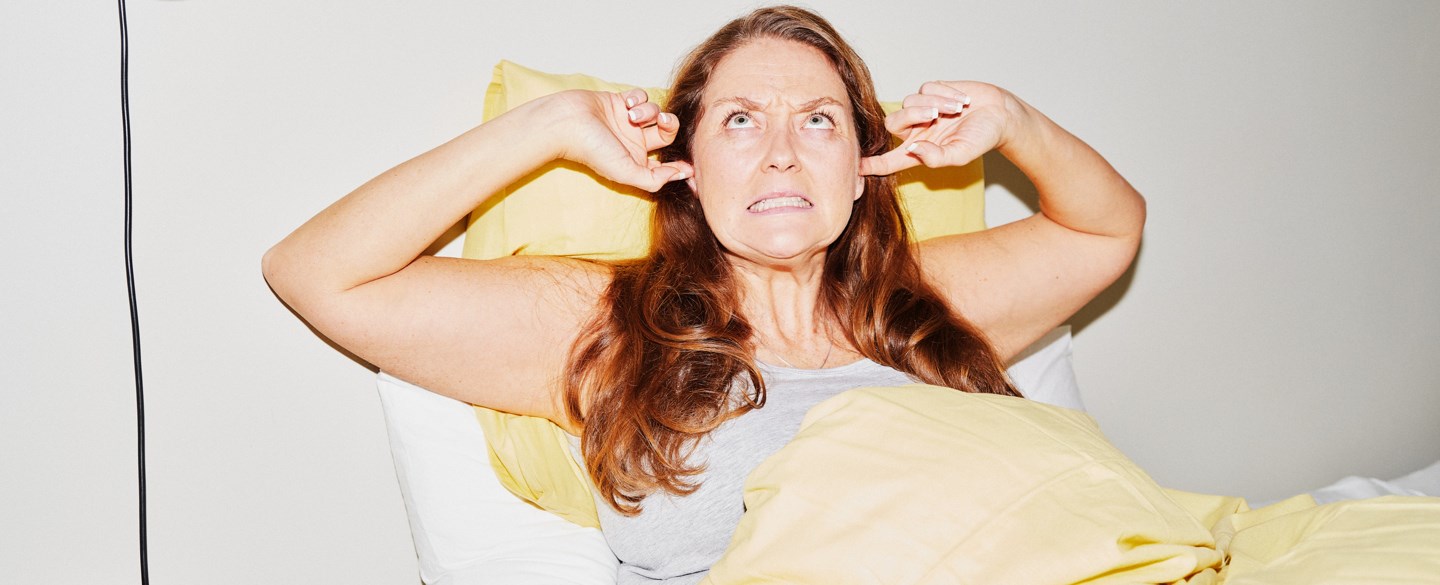 En kvinna sitter i sängen och håller för öronen.