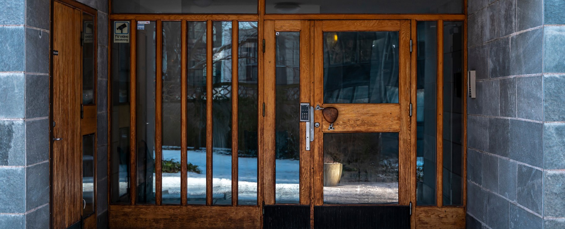 Portingång. Dörr- och fönsterglas med bruna lister av trä. Grå kakelväggar på sidorna.