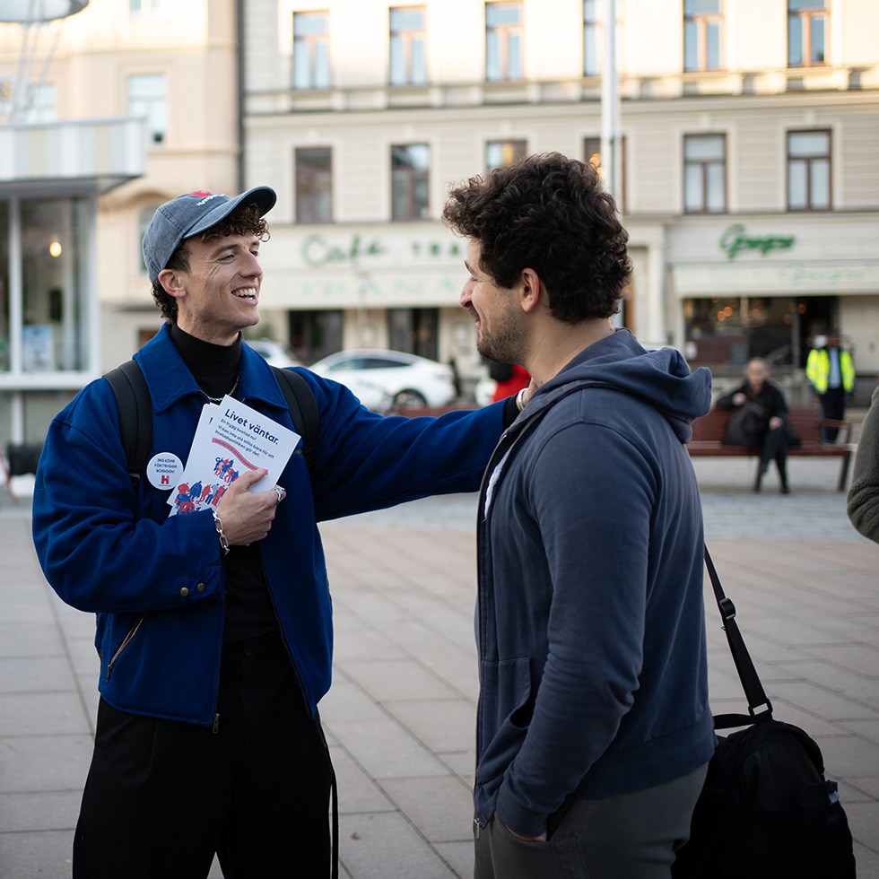 Foto på Ola Palmgren som står och pratar med en kille. I handen har han en flyer och på jackan sitter en pin med Hyresgästföreningens logga.