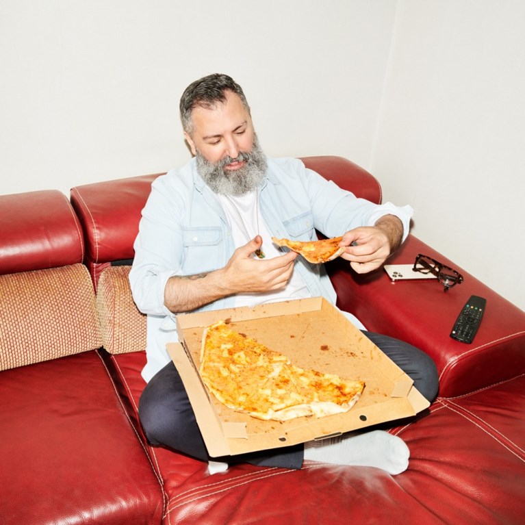 En man sitter i en soffa och äter pizza direkt ur kartongen.