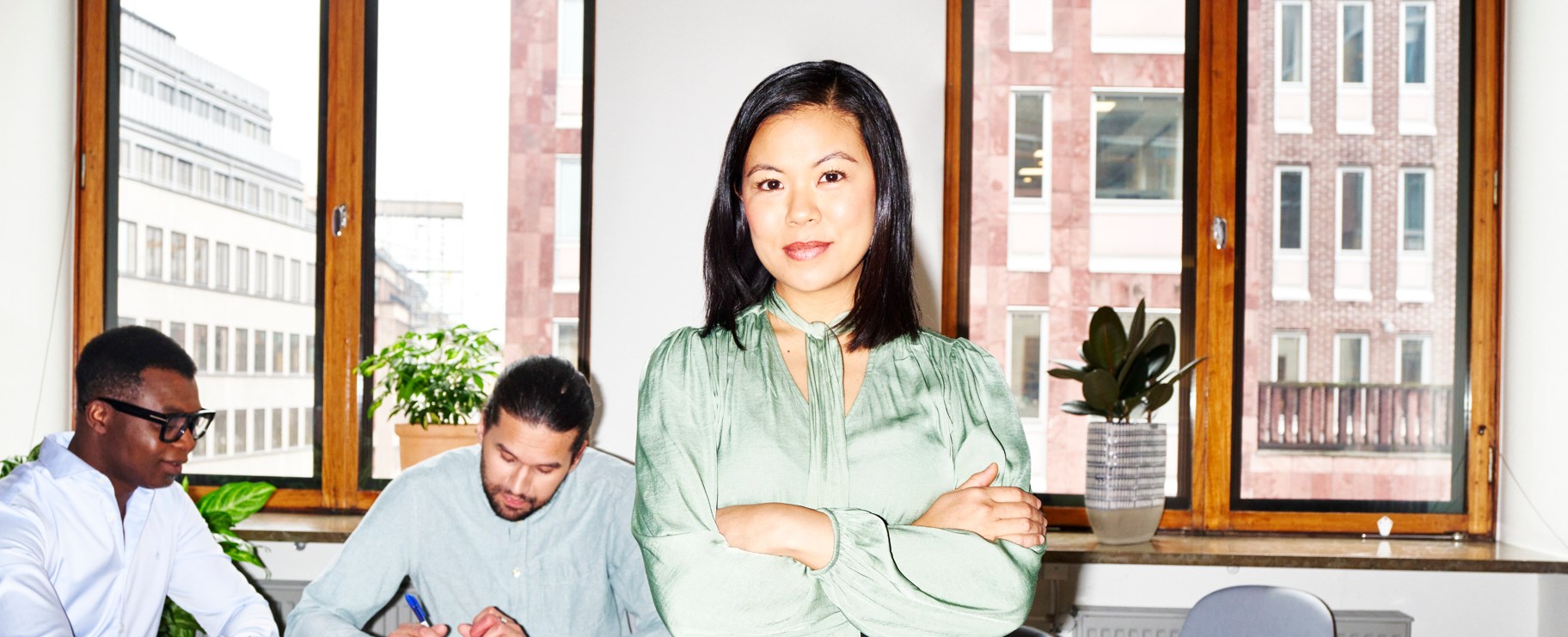 Kvinna på kontor med armarna i kors och två män i bakgrunden